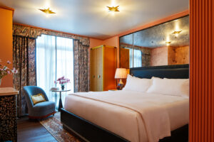 Tidily arranged bedroom with pristine bed, sofa with a table by the window, illuminated by warm golden light.