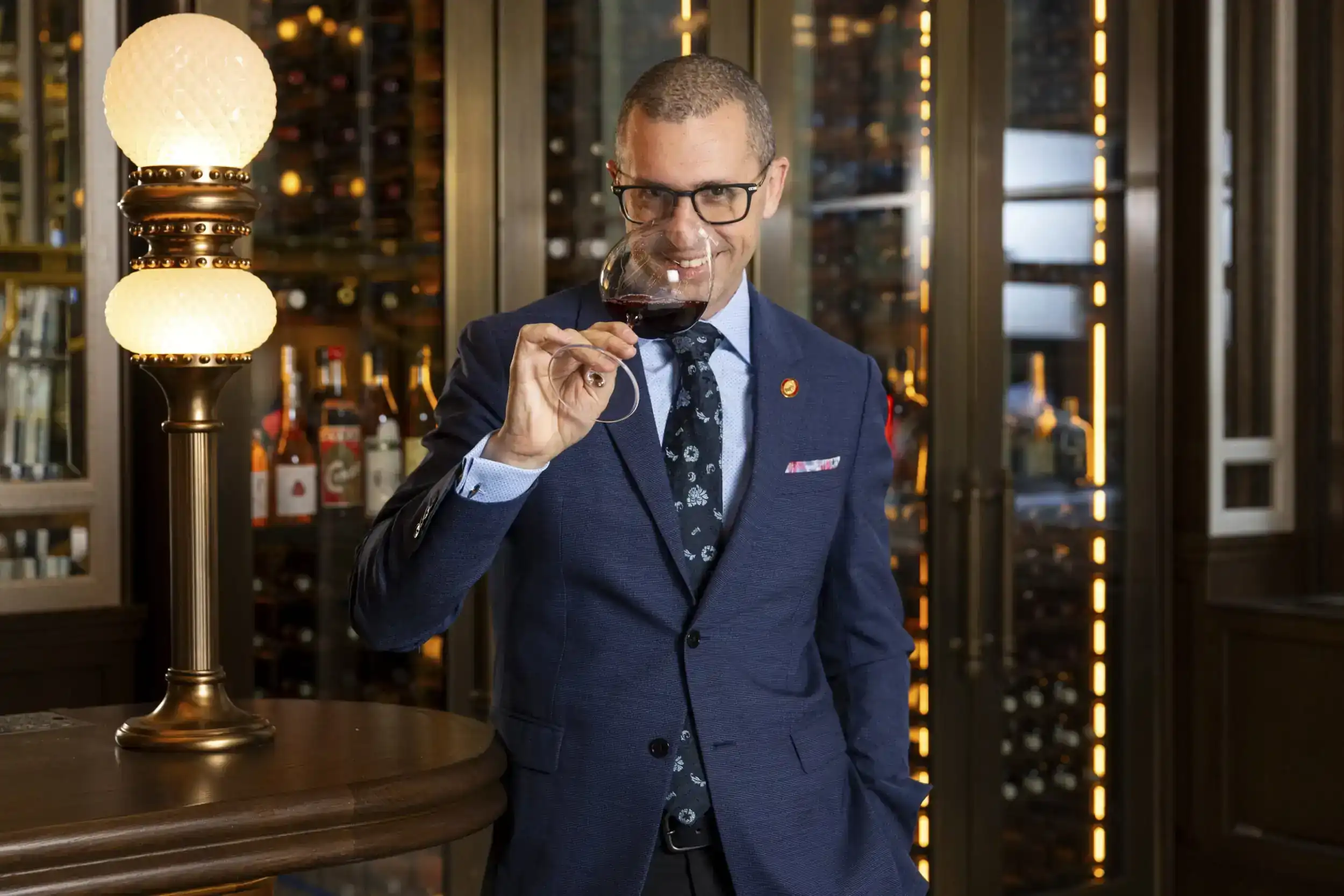 A man wearing a blue suit, holding a wine glass near a table adorned with a table lamp.