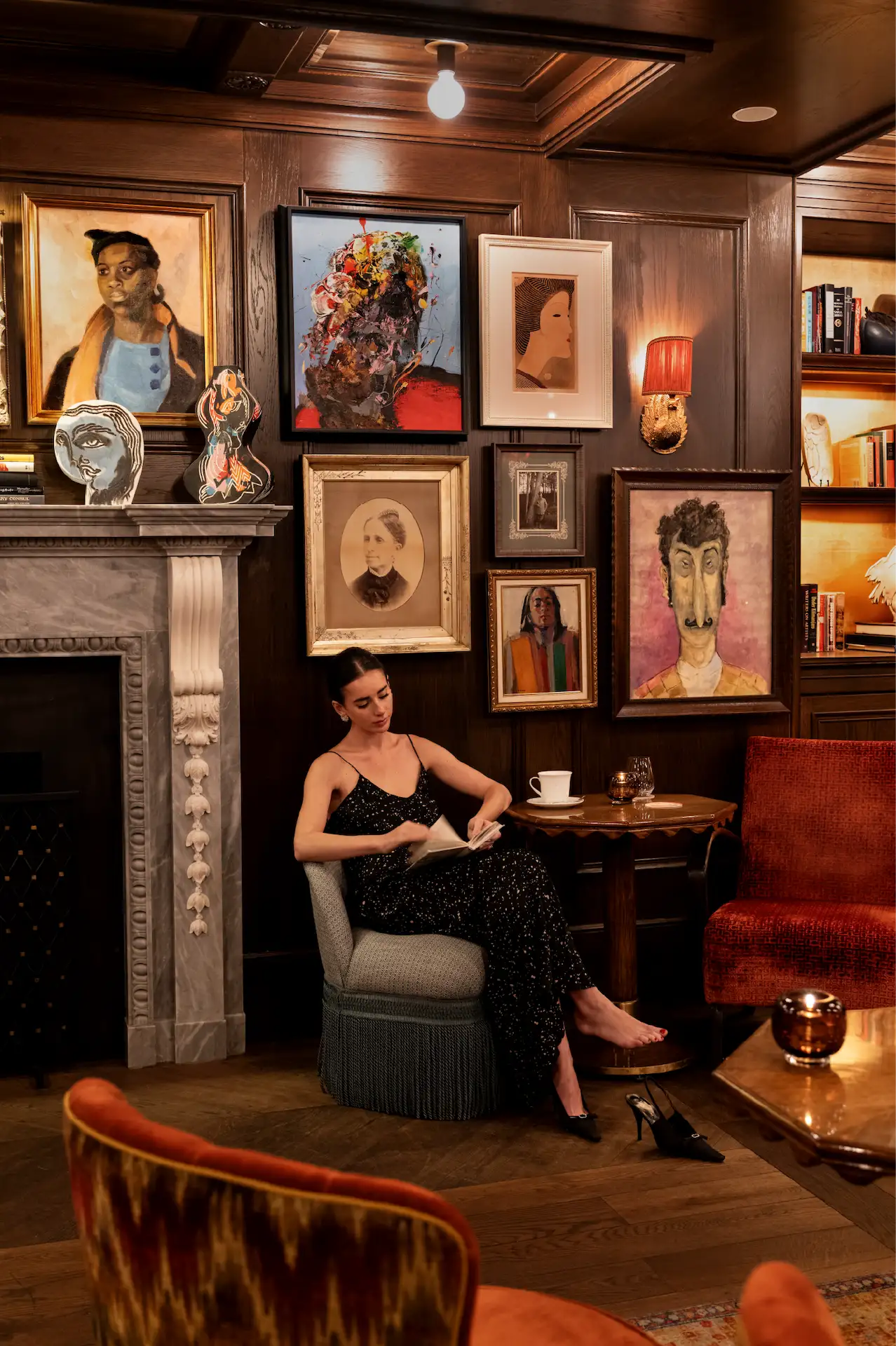 Woman in black sparkly dress reading in art-filled lounge with marble fireplace, eclectic portraits & warm ambient lighting.