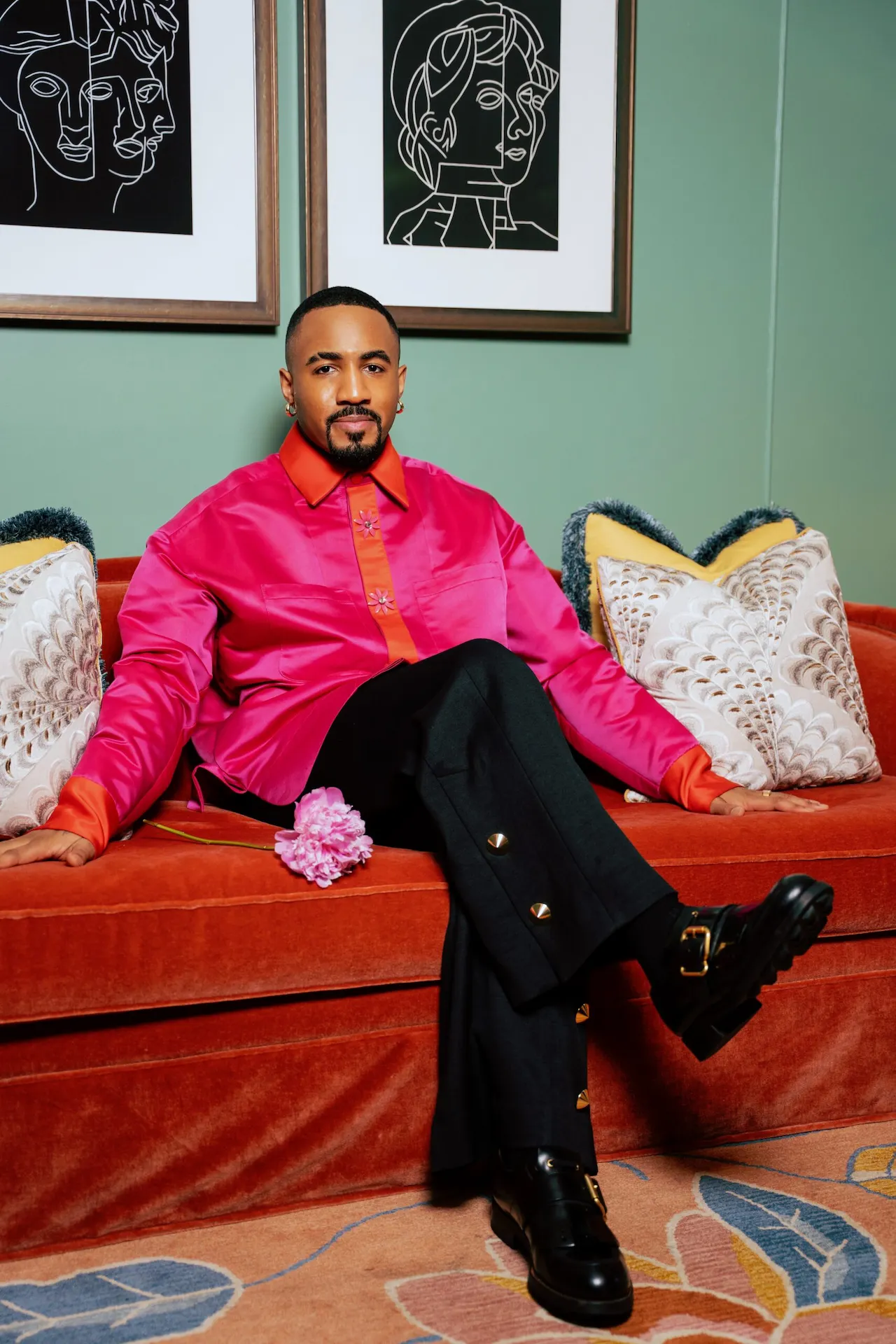 Person in vibrant pink and red embroidered shirt with black pants and gold buttons sitting on rust velvet couch.