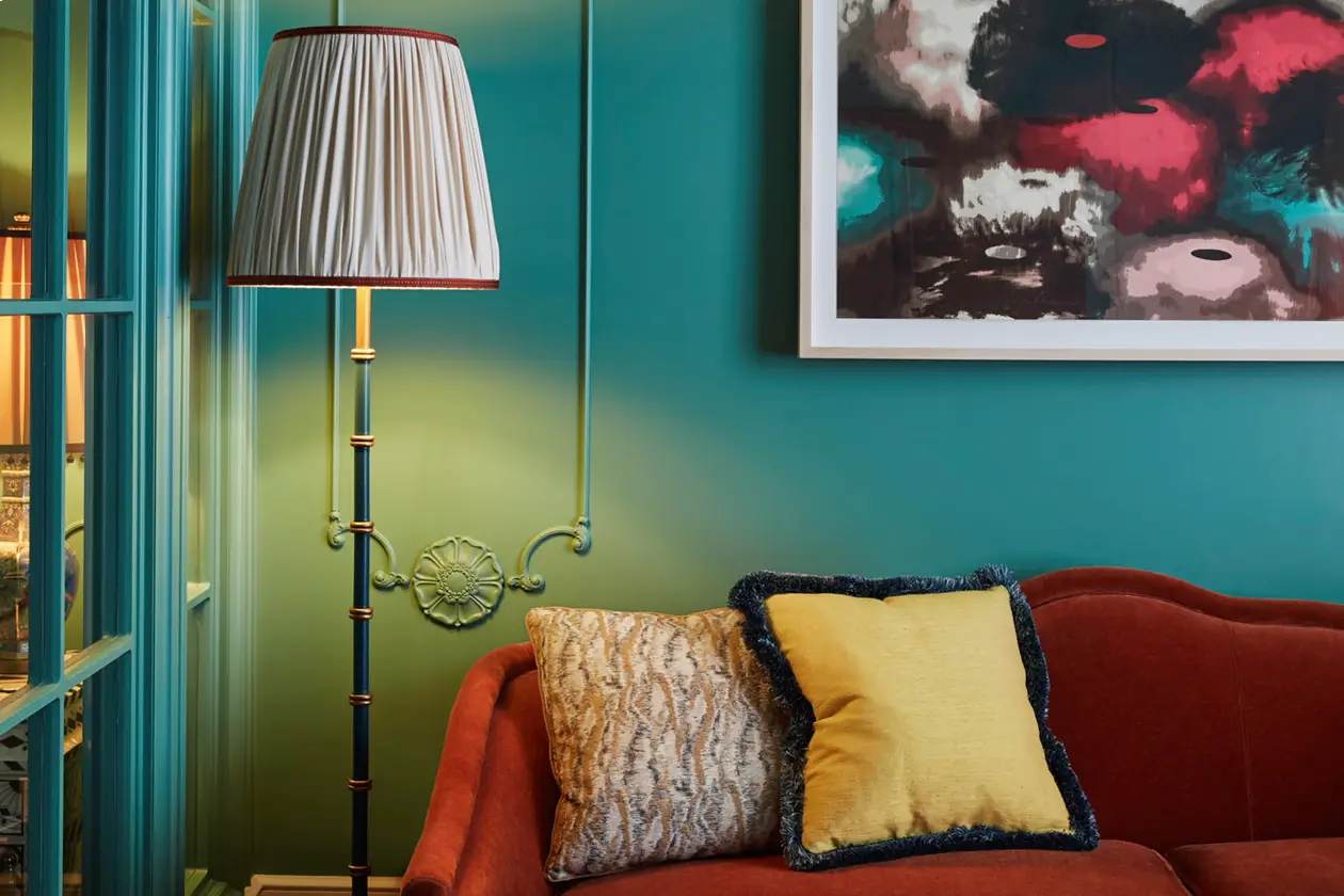 Red velvet sofa with beige and yellow pillows against teal wall, framed abstract art and brass floor lamp with pleated shade.