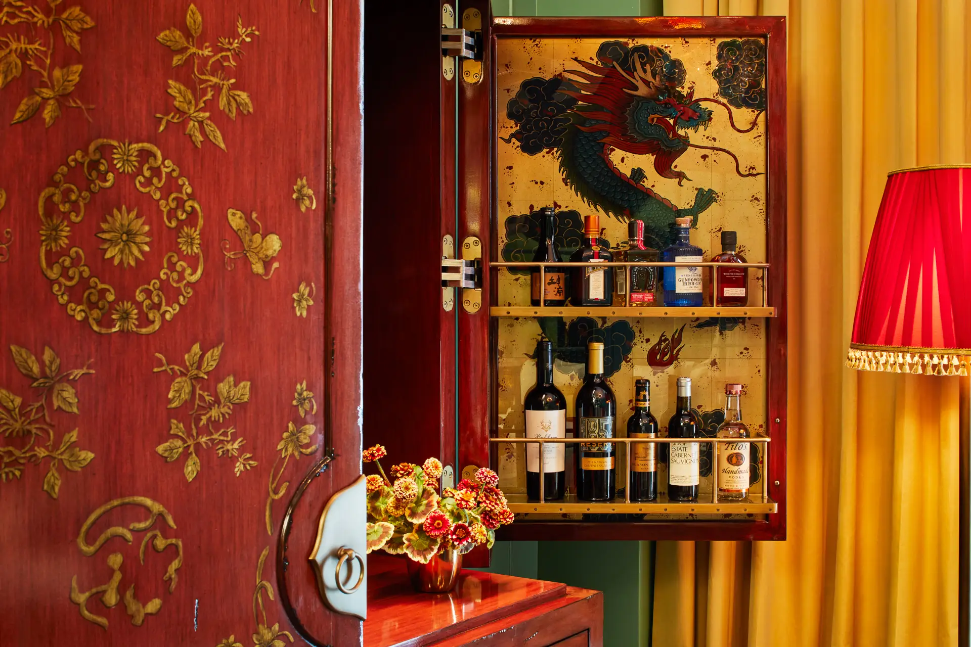 Ornate red and gold cabinet with dragon painting inside, stocked with bottles and flanked by lamp and floral arrangement.
