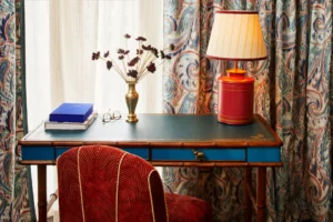 Elegant hotel desk with blue surface, red lamp & patterned chair, set against vibrant curtains and cozy decor.