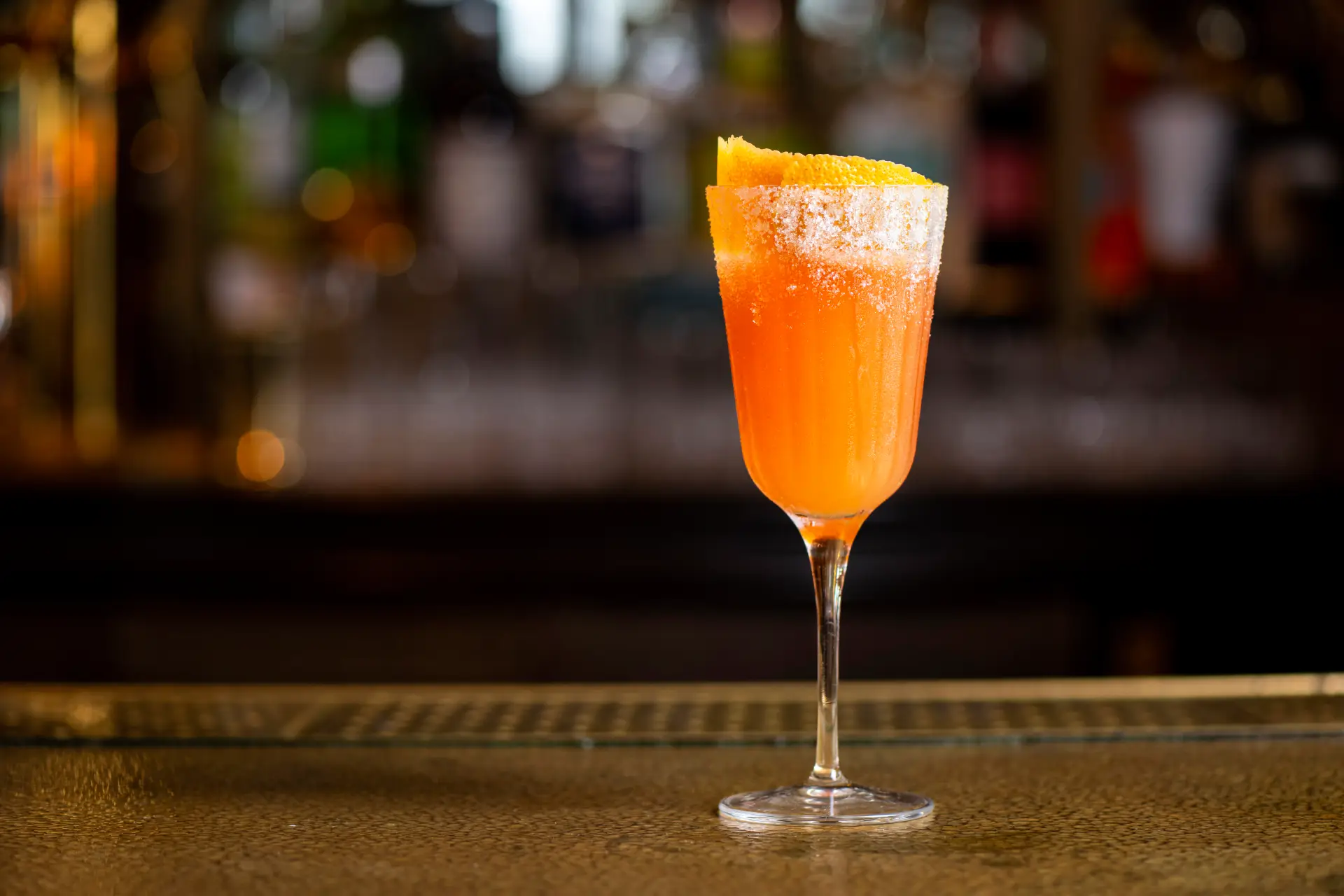 Orange cocktail in a sugar-rimmed glass with citrus peel garnish, set against a blurred background highlighting its vibrant color.