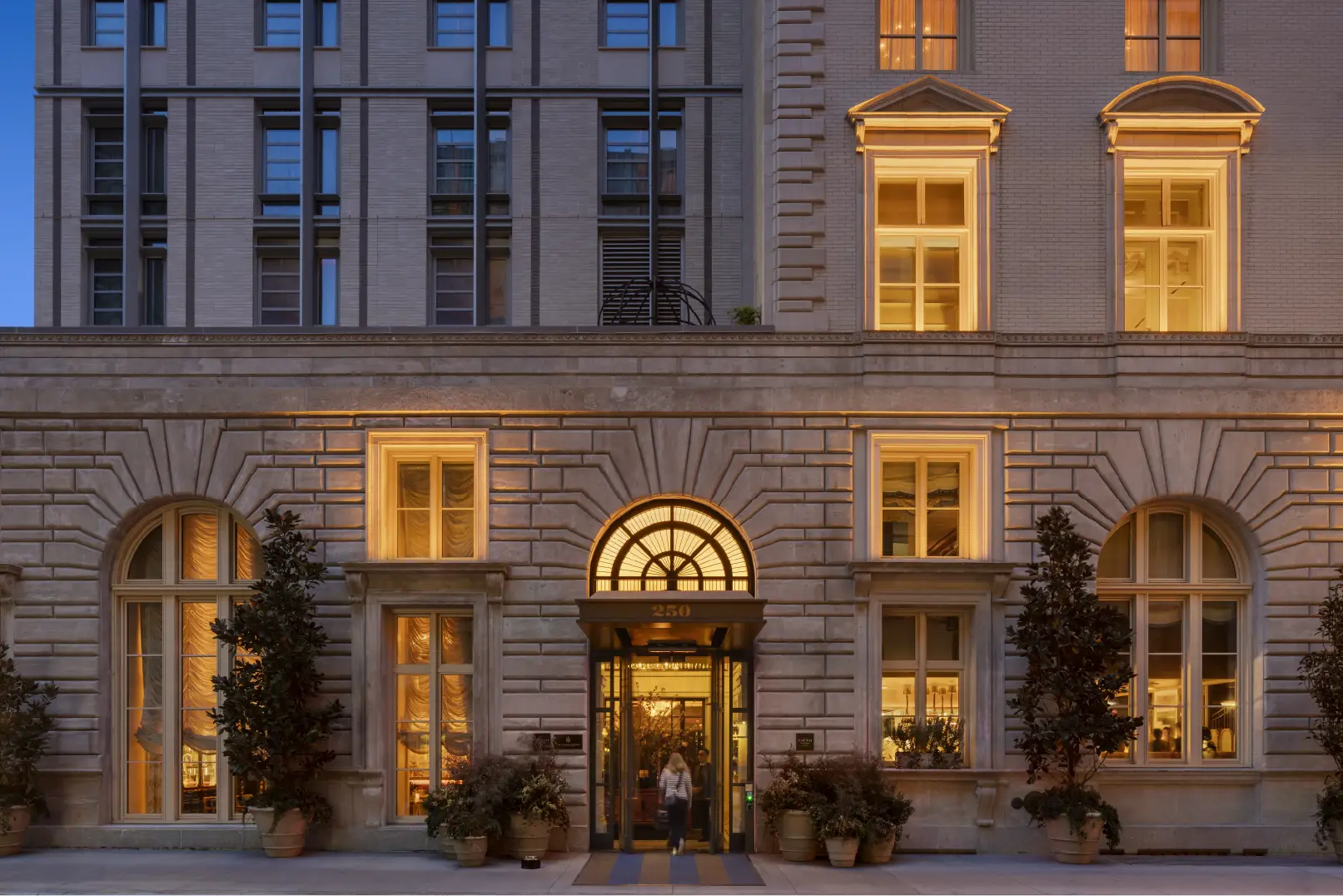 Elegant hotel entrance with arched window, stone facade, and warm lighting, framed by potted plants and classical detailing.