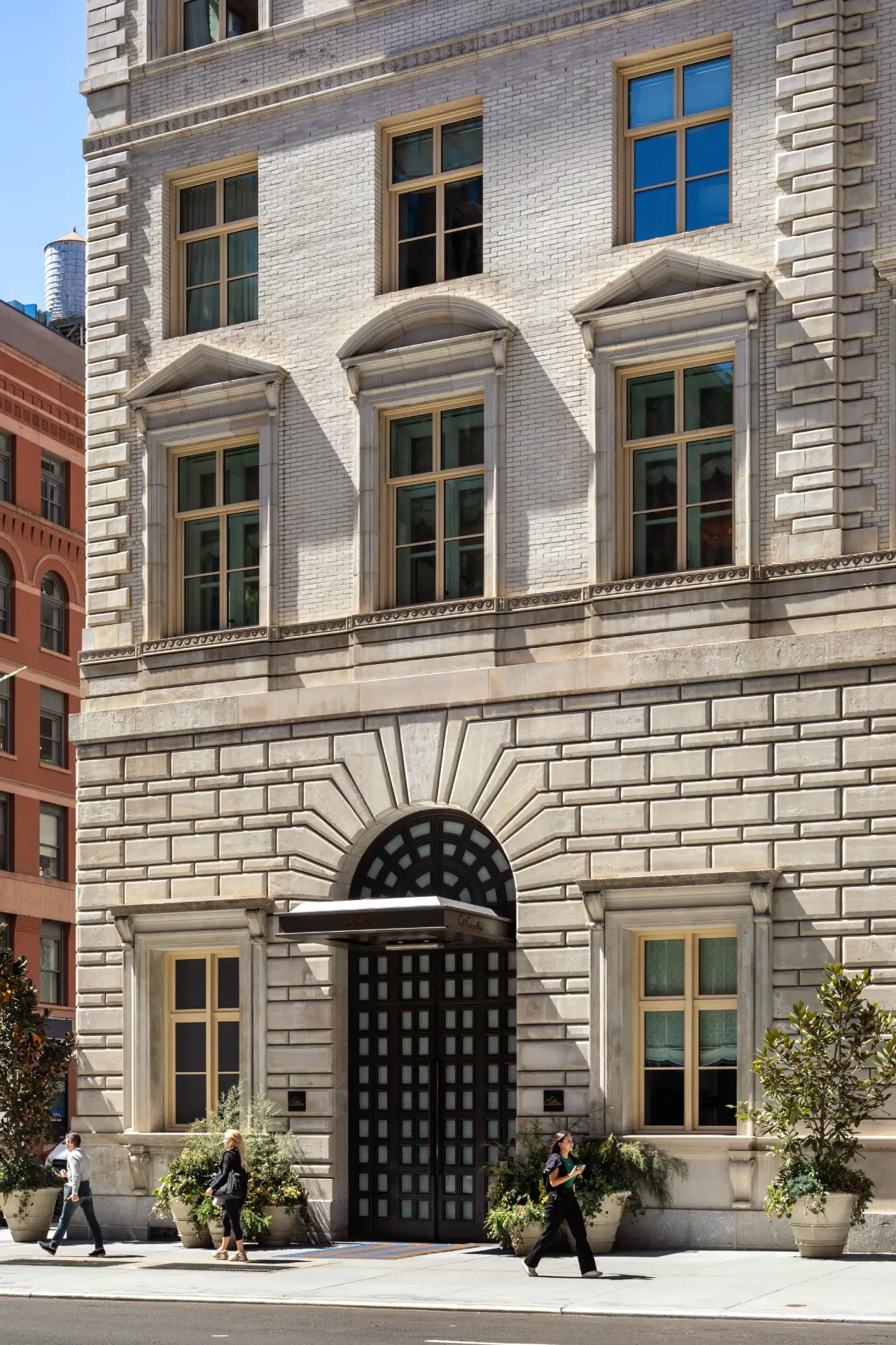 Classical stone building facade with arched doorway, decorative masonry, symmetrical windows, plants & people walking by