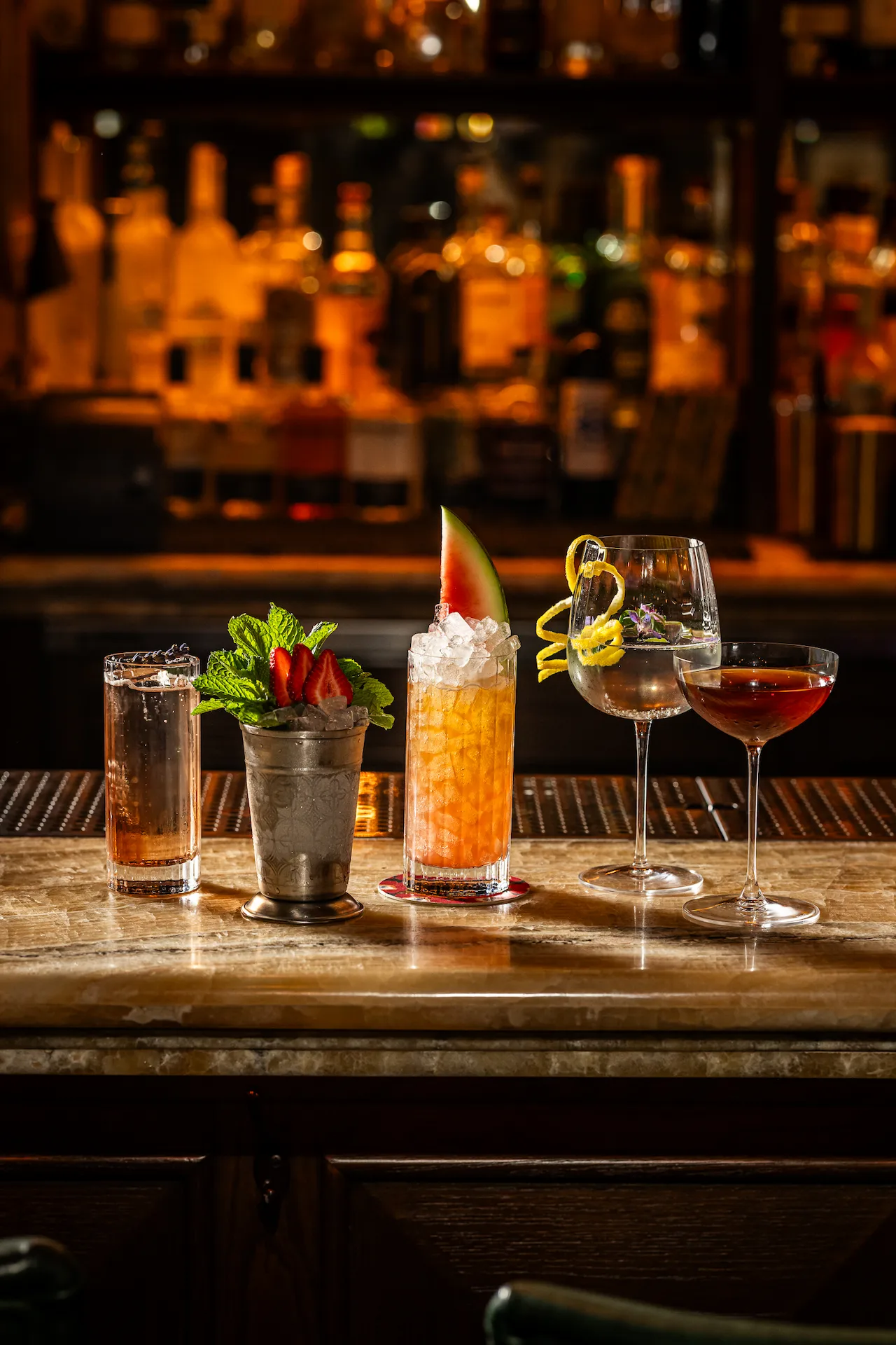 Five colorful cocktails on marble bar, each uniquely garnished, set against a warm, inviting backdrop of liquor bottles.
