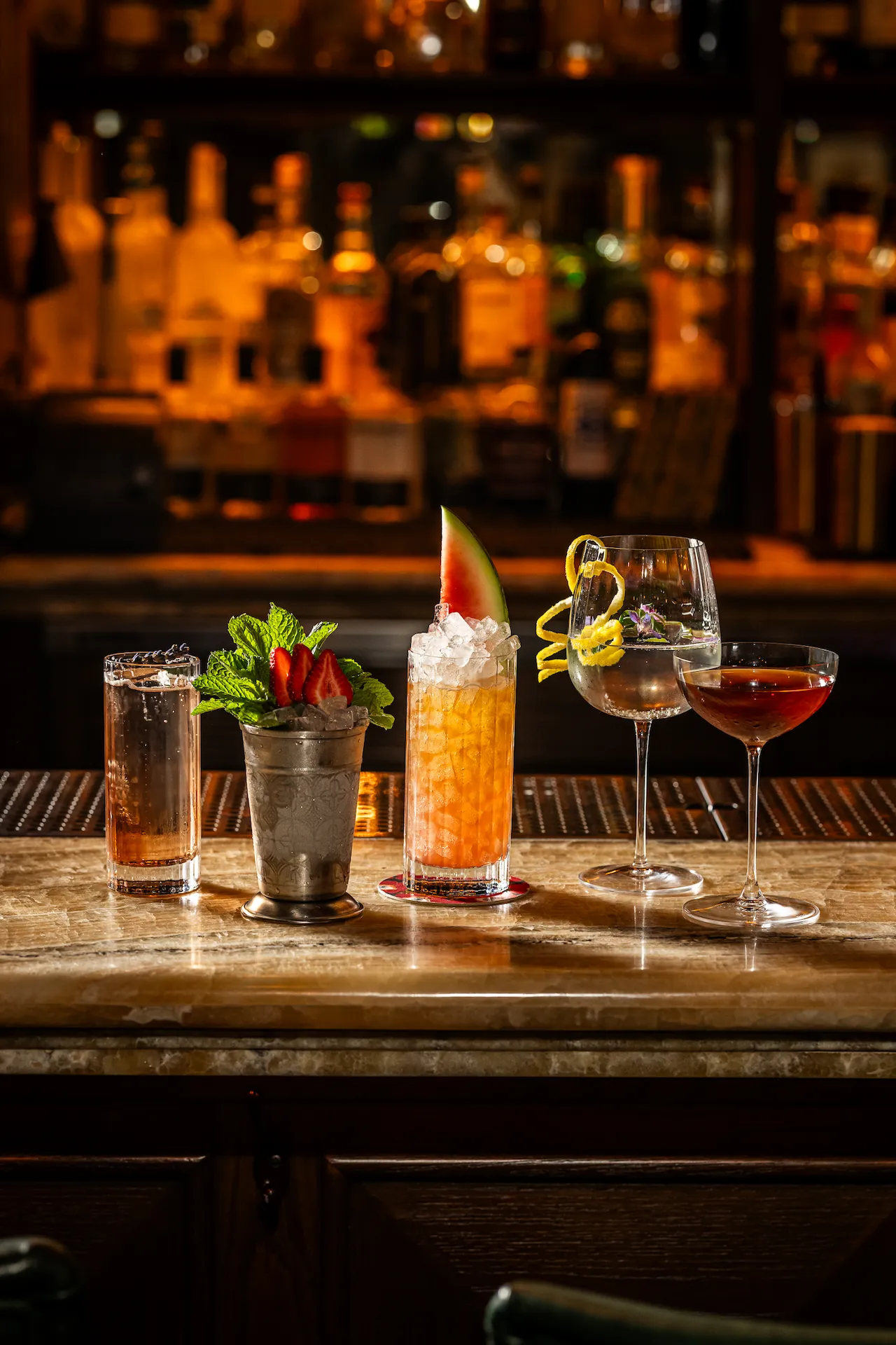 Five colorful cocktails on marble bar, each uniquely garnished, set against a warm, inviting backdrop of liquor bottles.