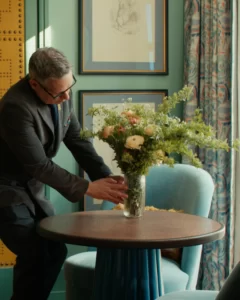 Person in dark suit adjusting floral arrangement on round table in stylish room with teal walls