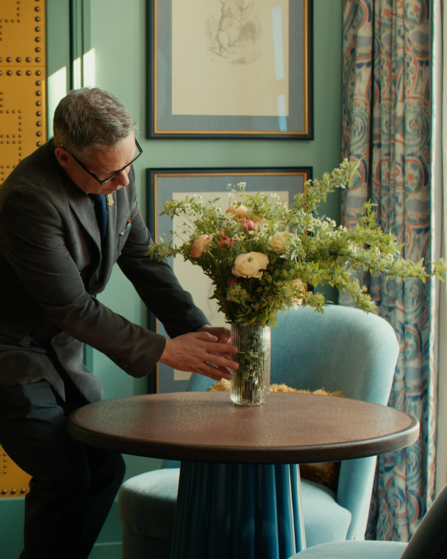 Person in dark suit adjusting floral arrangement on round table in stylish room with teal walls