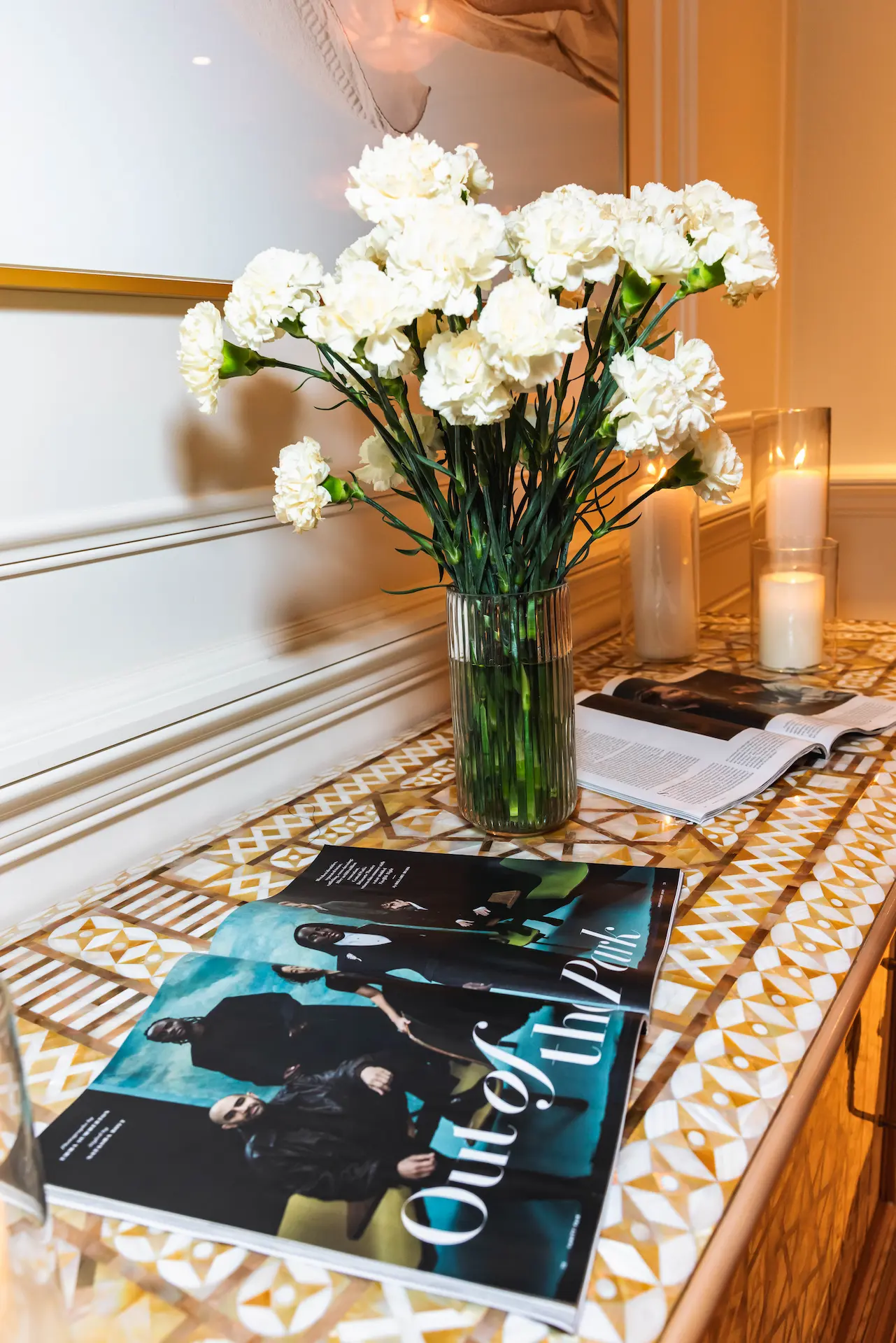Clear glass vase with white carnations on patterned console table beside magazines, open book, lit candle & framed artwork.