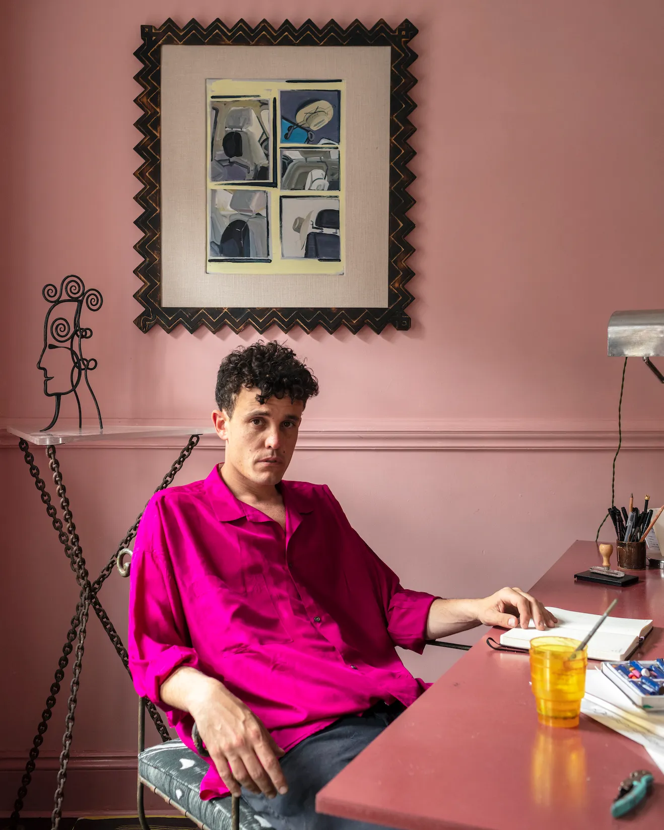 Person in bright pink shirt sits at desk with yellow glass and notebook, framed by pink walls and abstract zigzag artwork.