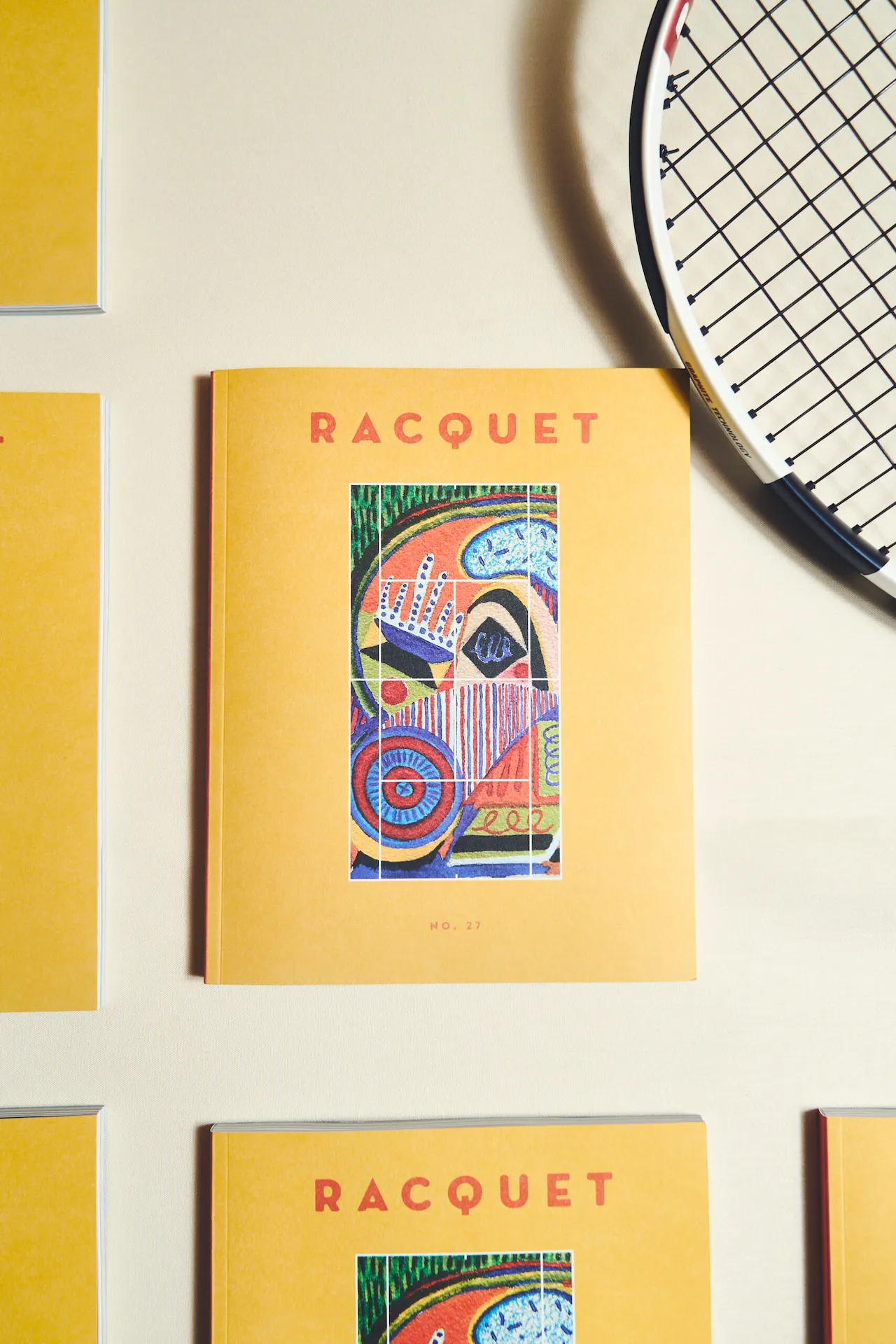 Several copies of Racquet magazine No. 17 with yellow covers and tennis-themed artwork arranged beside a racket.