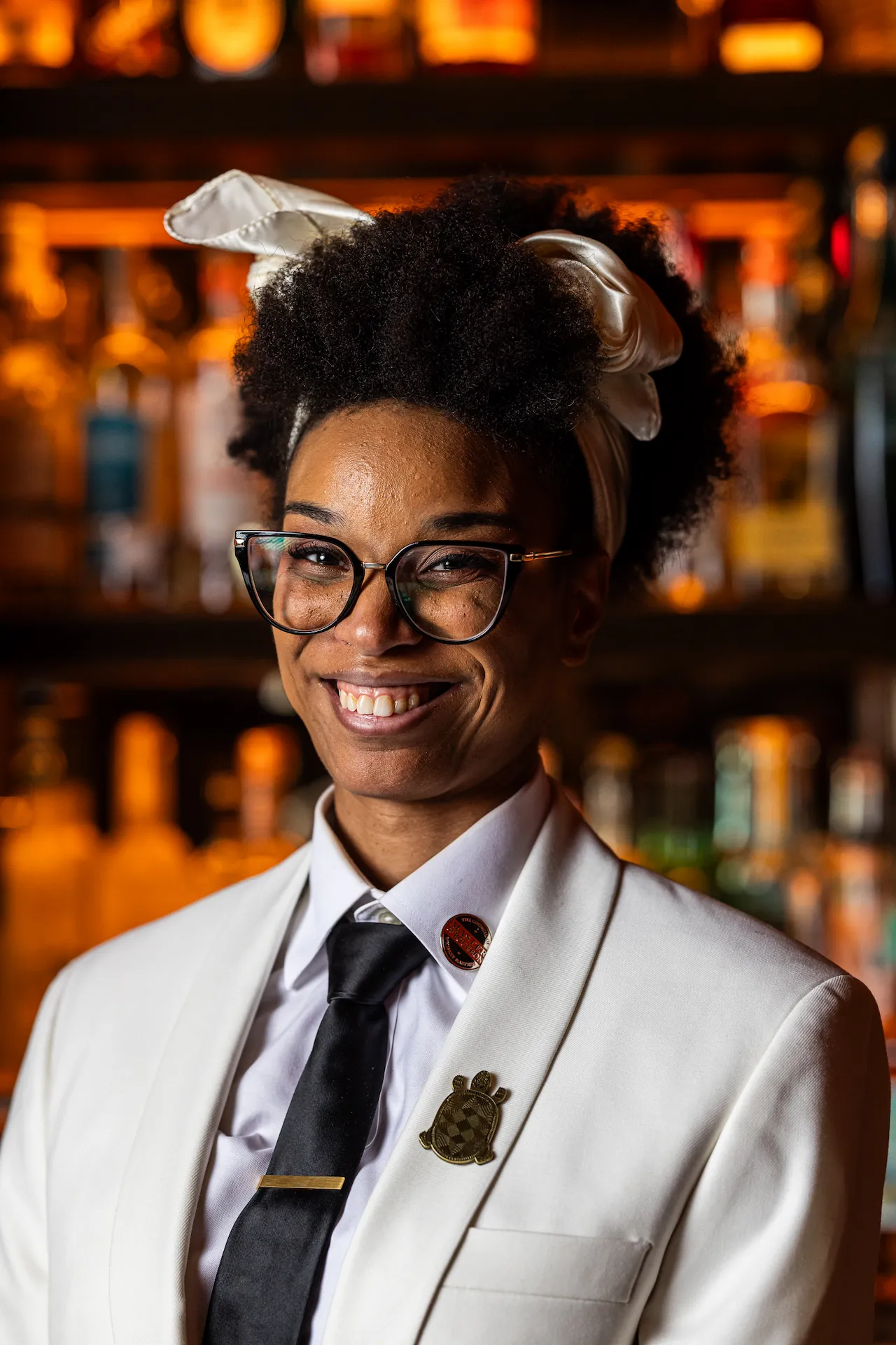 Person in white suit with gold tie clip and satin headband standing in front of warmly lit bar shelf with bottles.