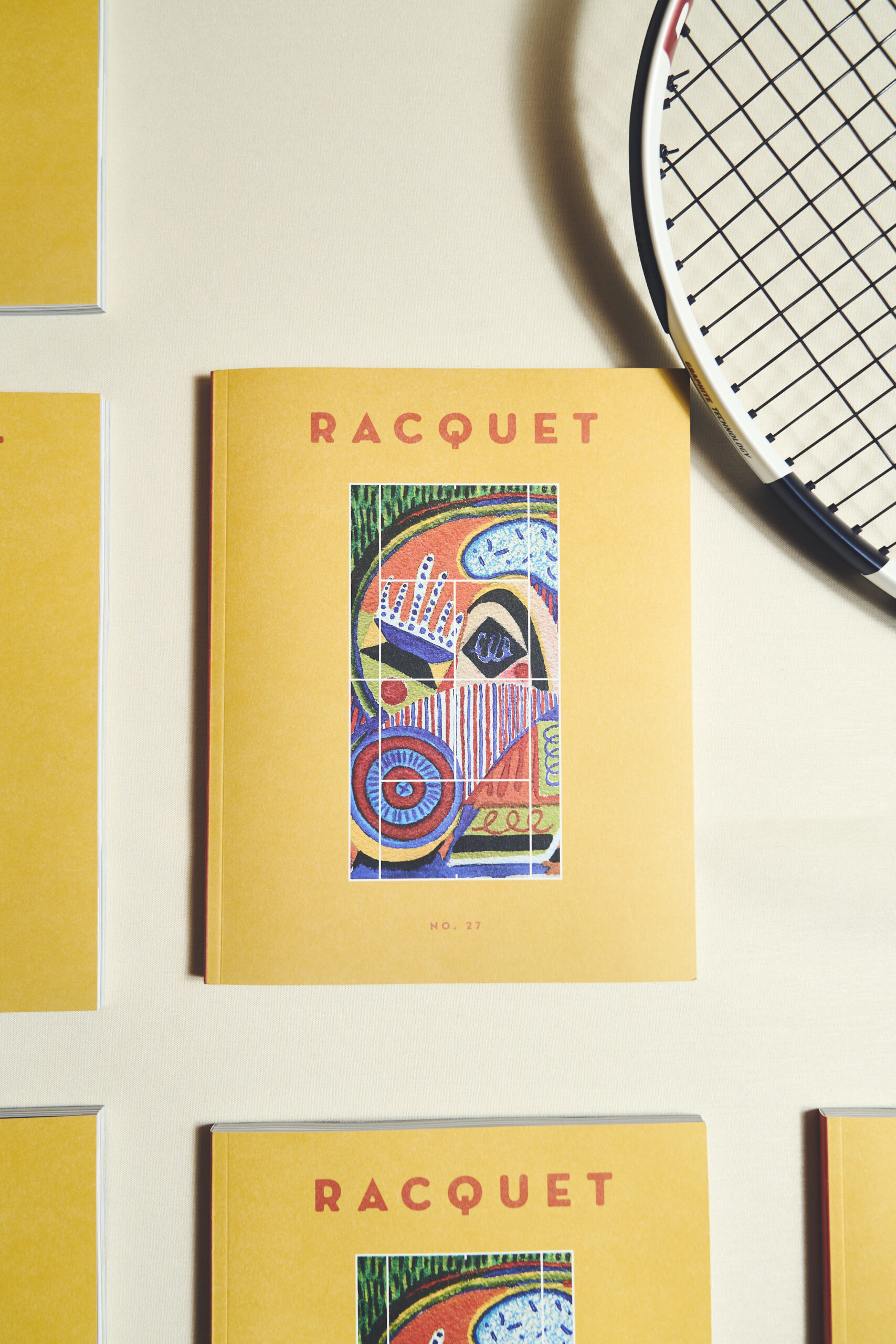 Several copies of Racquet magazine No. 17 with yellow covers and tennis-themed artwork arranged beside a racket.