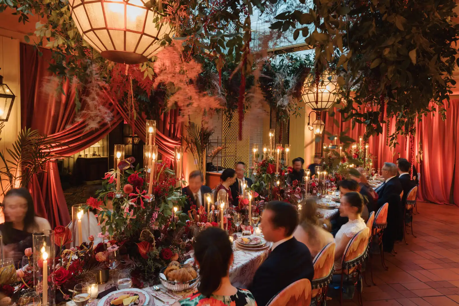 Elegant dinner party with floral centerpieces, hanging lanterns, red drapery, and guests in formal attire.