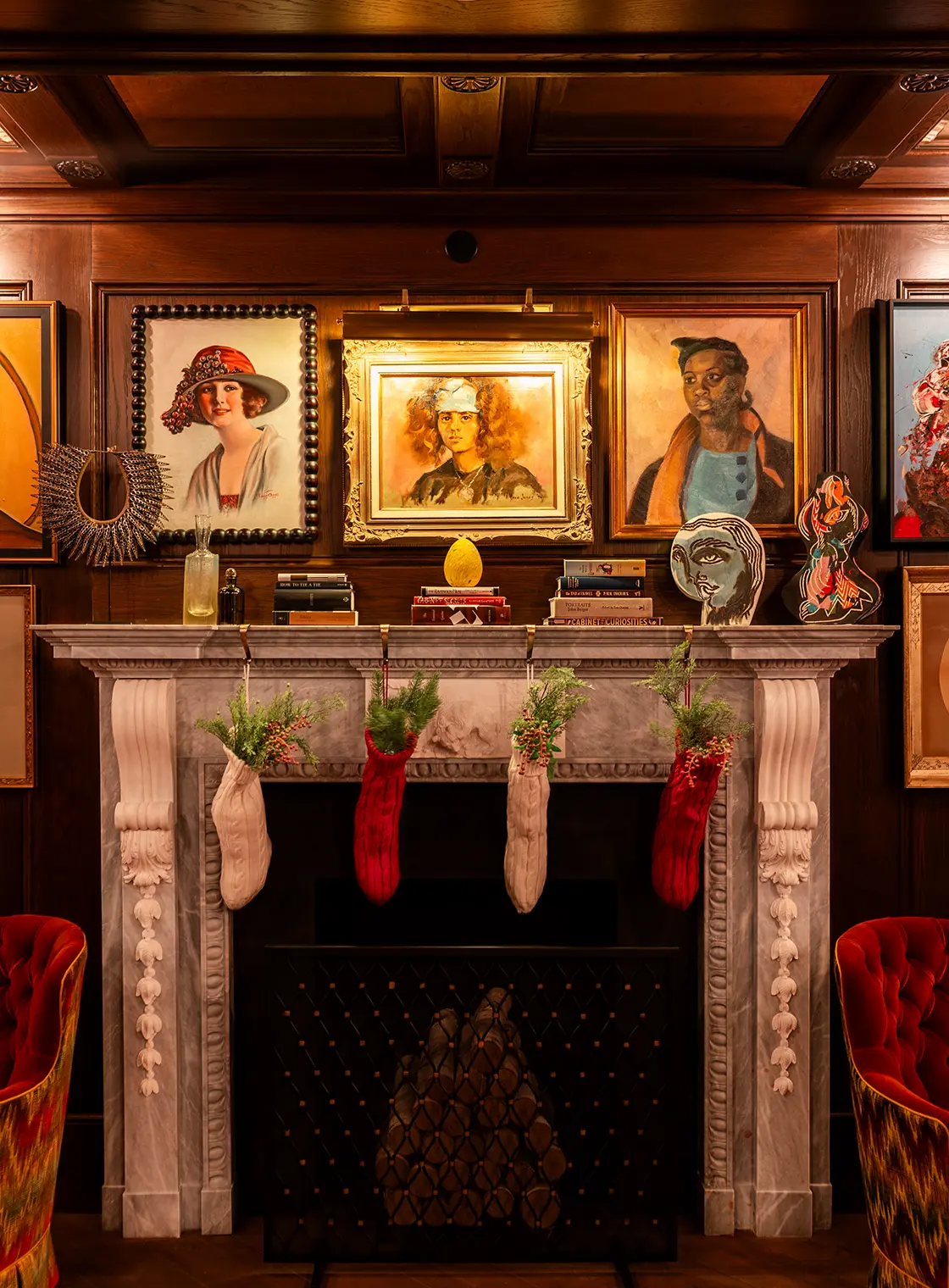 Ornate interior with marble fireplace, festive stockings, eclectic artwork, and plush red seating.