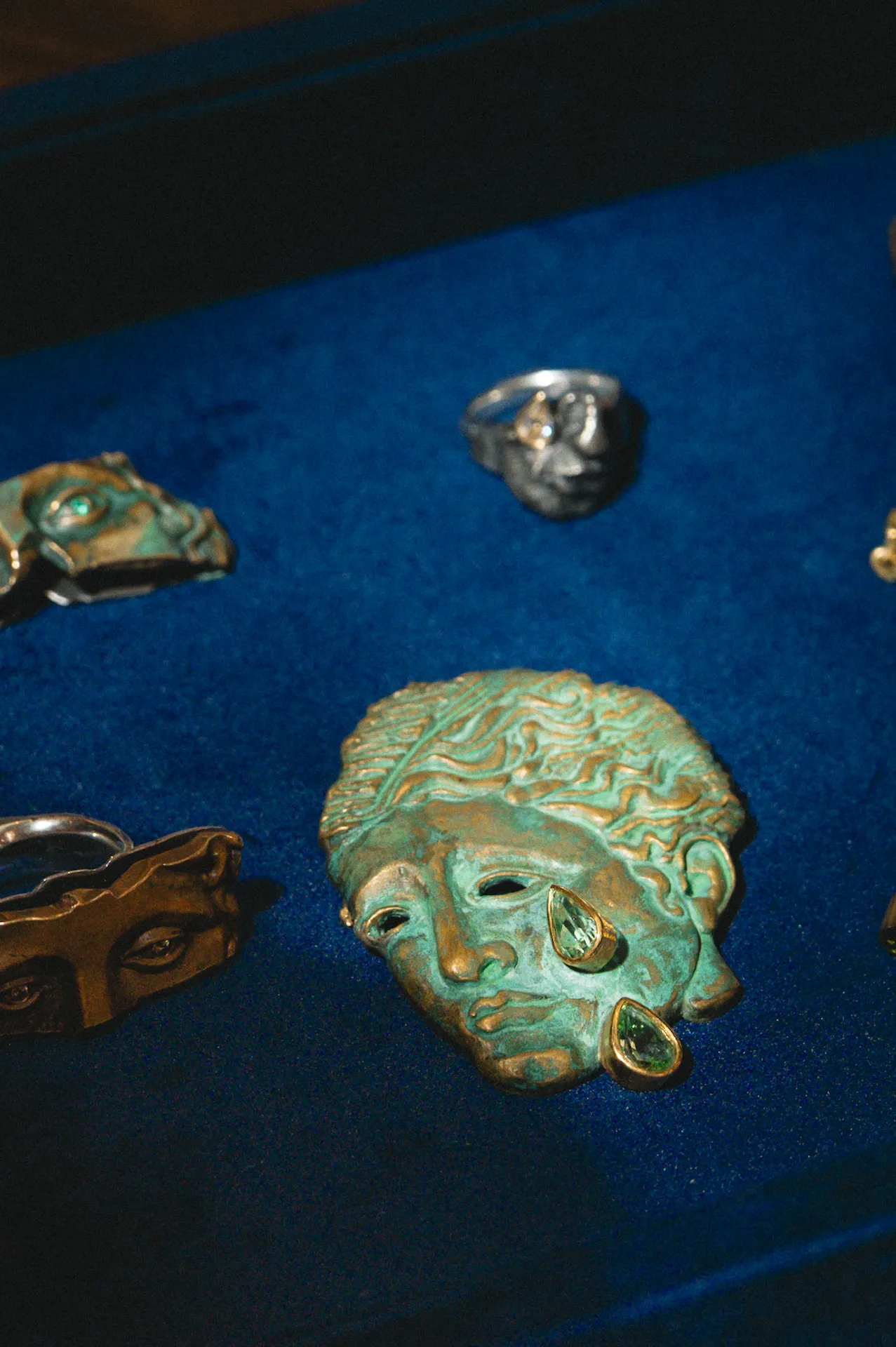 Artistic jewelry display on blue velvet featuring sculpted face brooch with green gemstone tears and metallic rings.