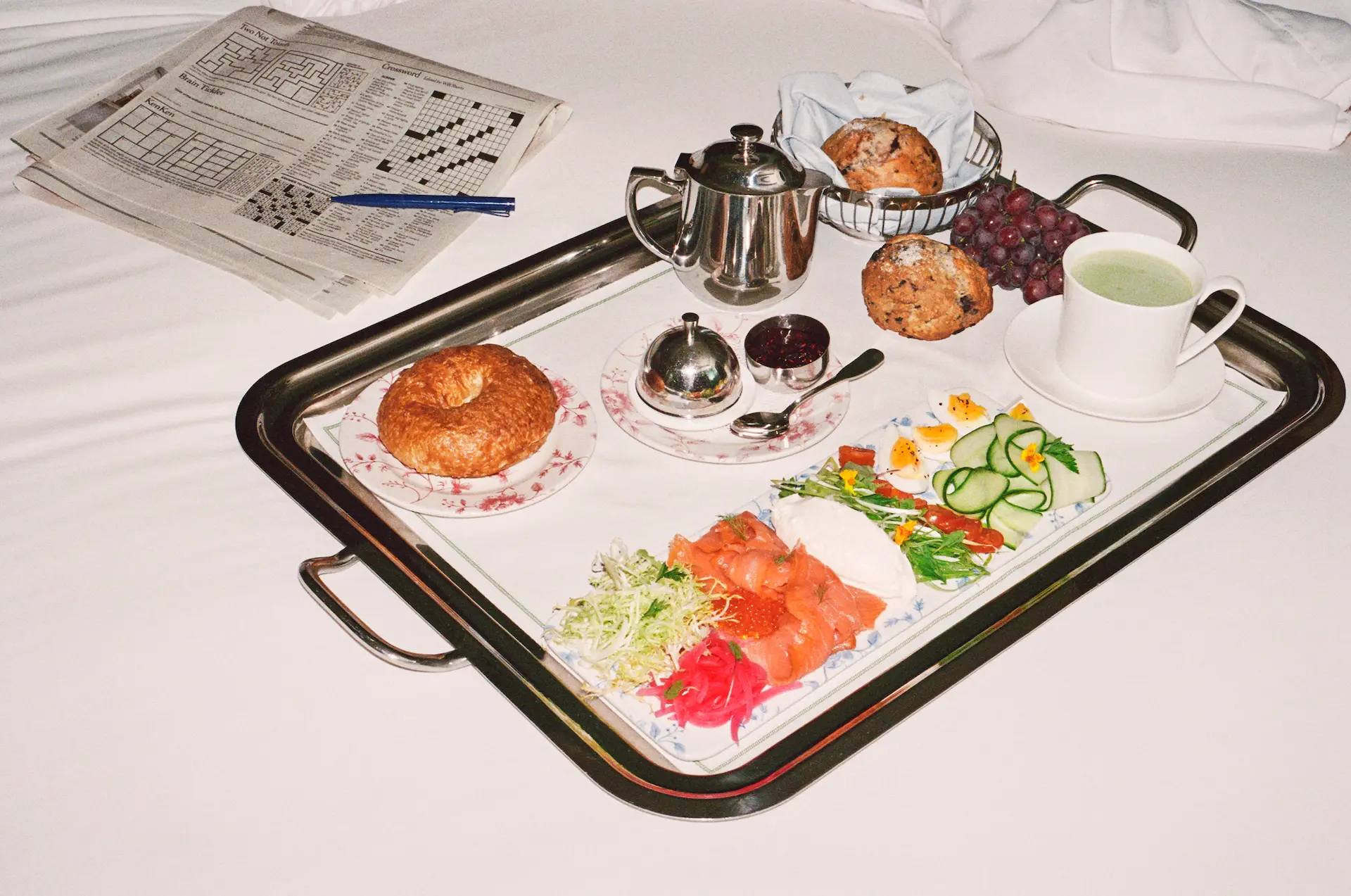 Breakfast tray on white bed with smoked salmon, tea, pastries, and newspaper, evoking a relaxed luxury morning moment.