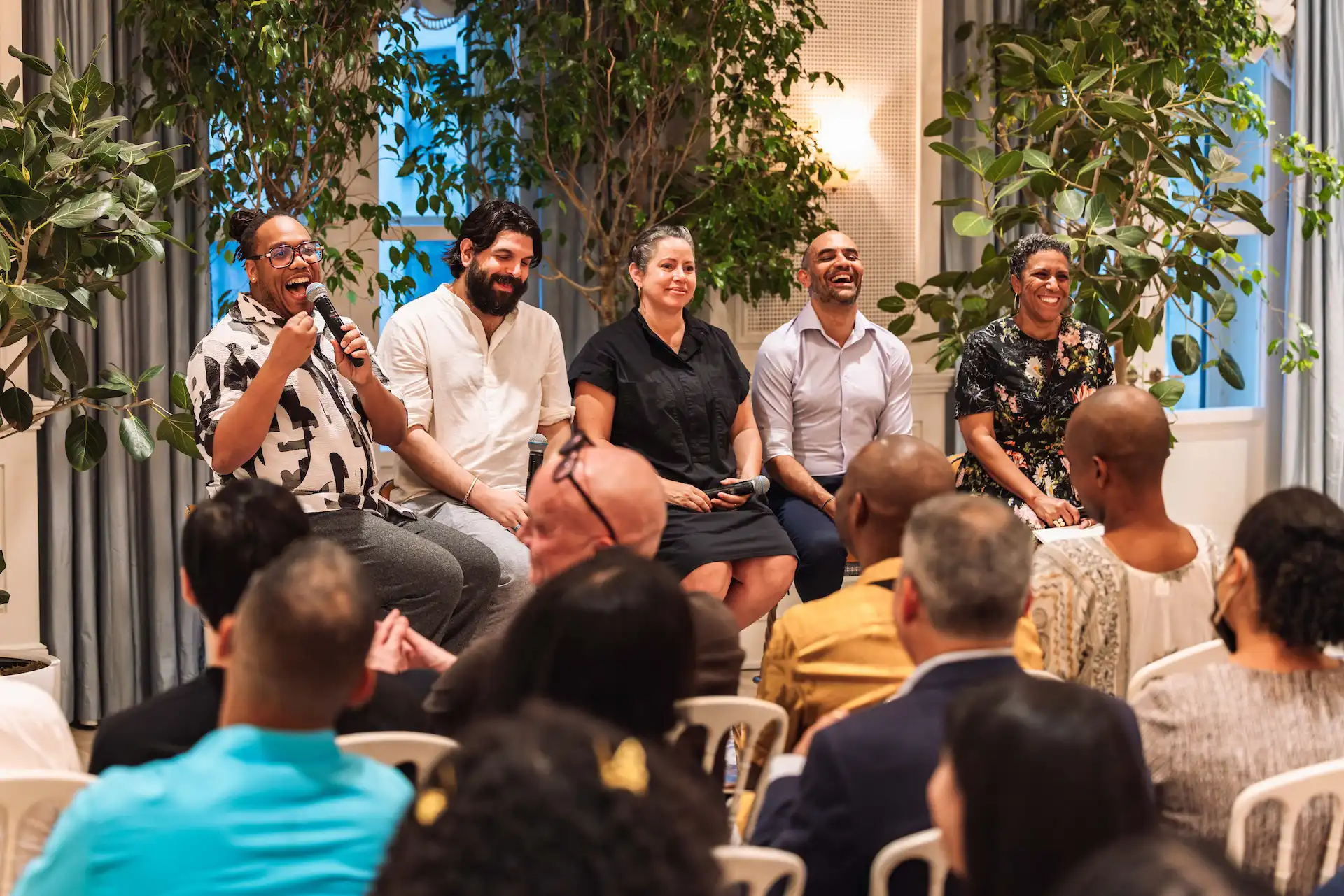 Panel discussion with five speakers seated in front of audience, surrounded by greenery in a stylish indoor event space.