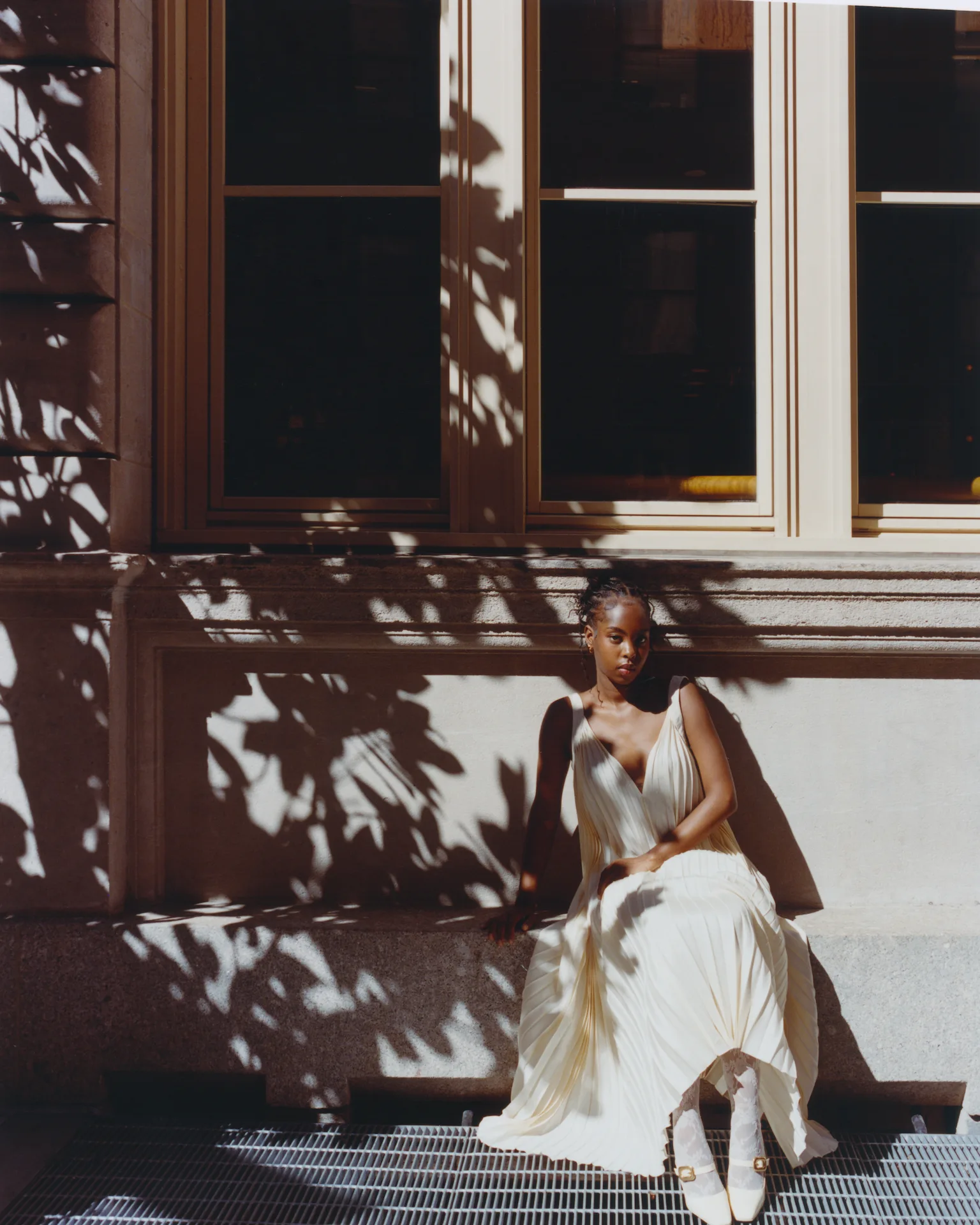 Person in flowing cream dress sits on stone bench, dappled in leafy shadows, evoking calm elegance outside a sunlit building.