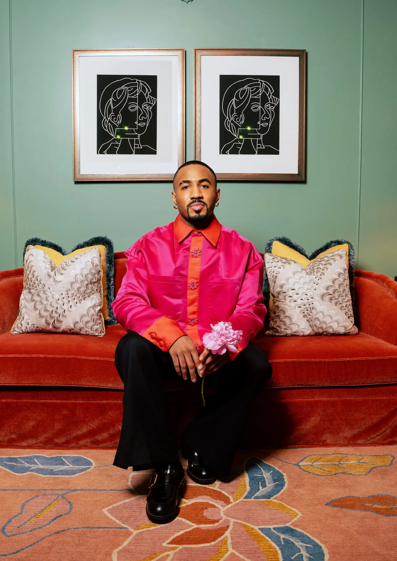 Person in bright pink shirt holding flower on red velvet couch, framed by green wall art and vibrant patterned rug below.
