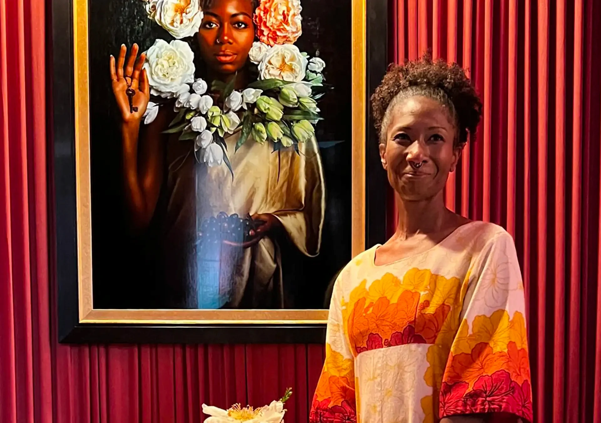 A lady with curly hair poses for a photo beside a beautiful portrait mounted on the wall.