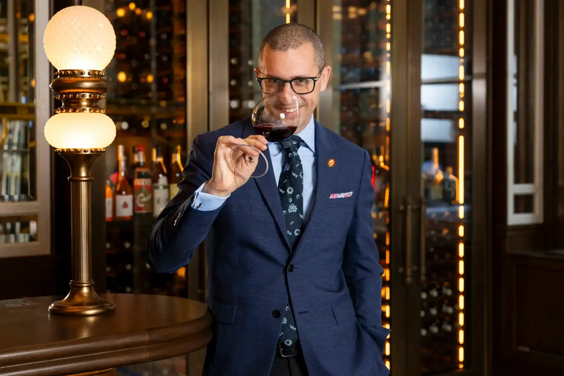 A man wearing a blue suit, holding a wine glass near a table adorned with a table lamp.