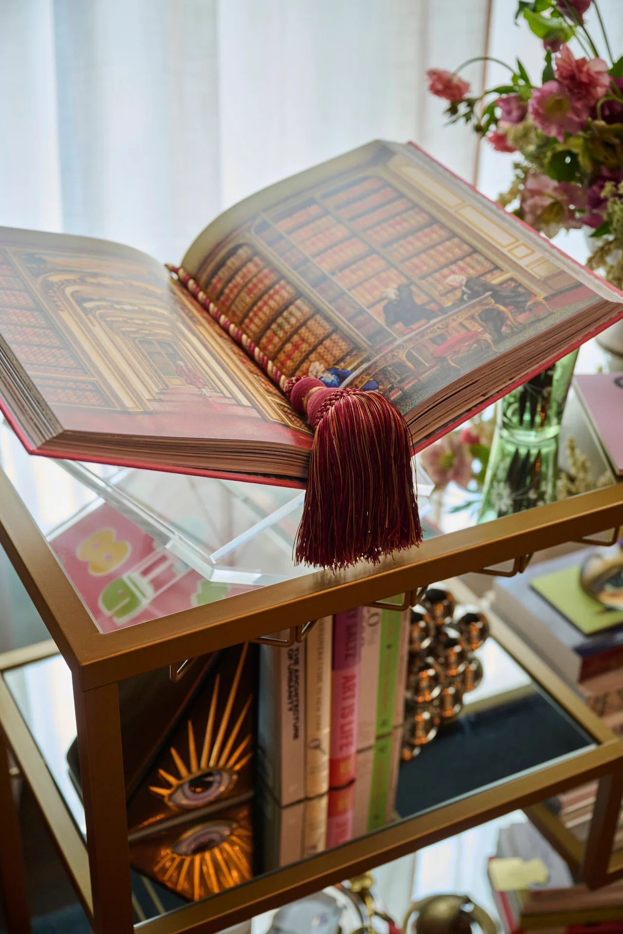 Open illustrated book on a glass shelf surrounded by flowers, colorful books, and decorative objects.