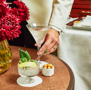 Hand holding a martini beside a table with garnishes, snacks, and red flowers at an elegant event.