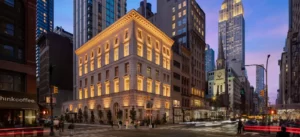 City street at dusk with the Empire State Building glowing above historic and modern buildings.