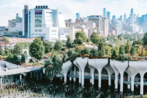 Little Island's sculpted concrete pilings and tree-lined park deck on the Hudson River, with the Whitney Museum and Manhattan skyline behind