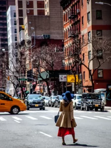 Woman in a camel trench coat, red midi skirt, and navy wide-brim hat crossing East 20th Street in Gramercy, Manhattan, beside a yellow NYC taxi