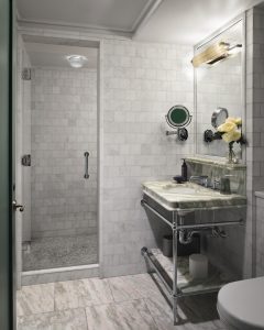 Marble-tiled bathroom with a glass shower enclosure on the left and a marble countertop vanity with a round sink on the right, topped by a vase of pale yellow roses and a wall mirror.