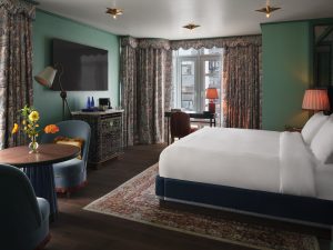 Hotel room with teal walls, large white bed, and patterned curtains; vintage cabinet, flowers on a round table, and warm lamps creating a cozy atmosphere in a bright windowed corner.