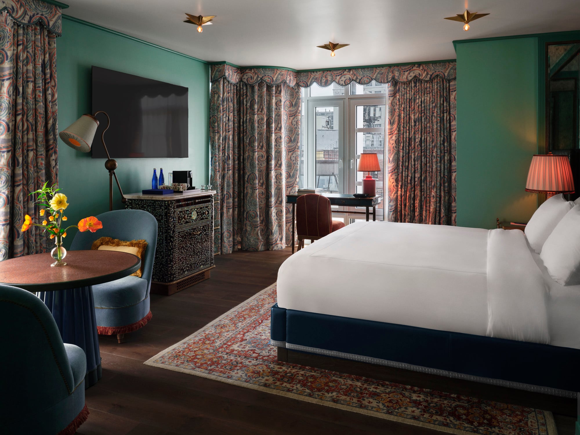 Hotel room with teal walls, large white bed, and patterned curtains; vintage cabinet, flowers on a round table, and warm lamps creating a cozy atmosphere in a bright windowed corner.