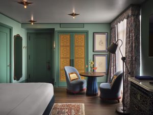 Teal living area with round wooden table, blue upholstered chairs, a vase of flowers, and patterned curtains beside a window. Green paneled doors and decorative panels are in the background.
