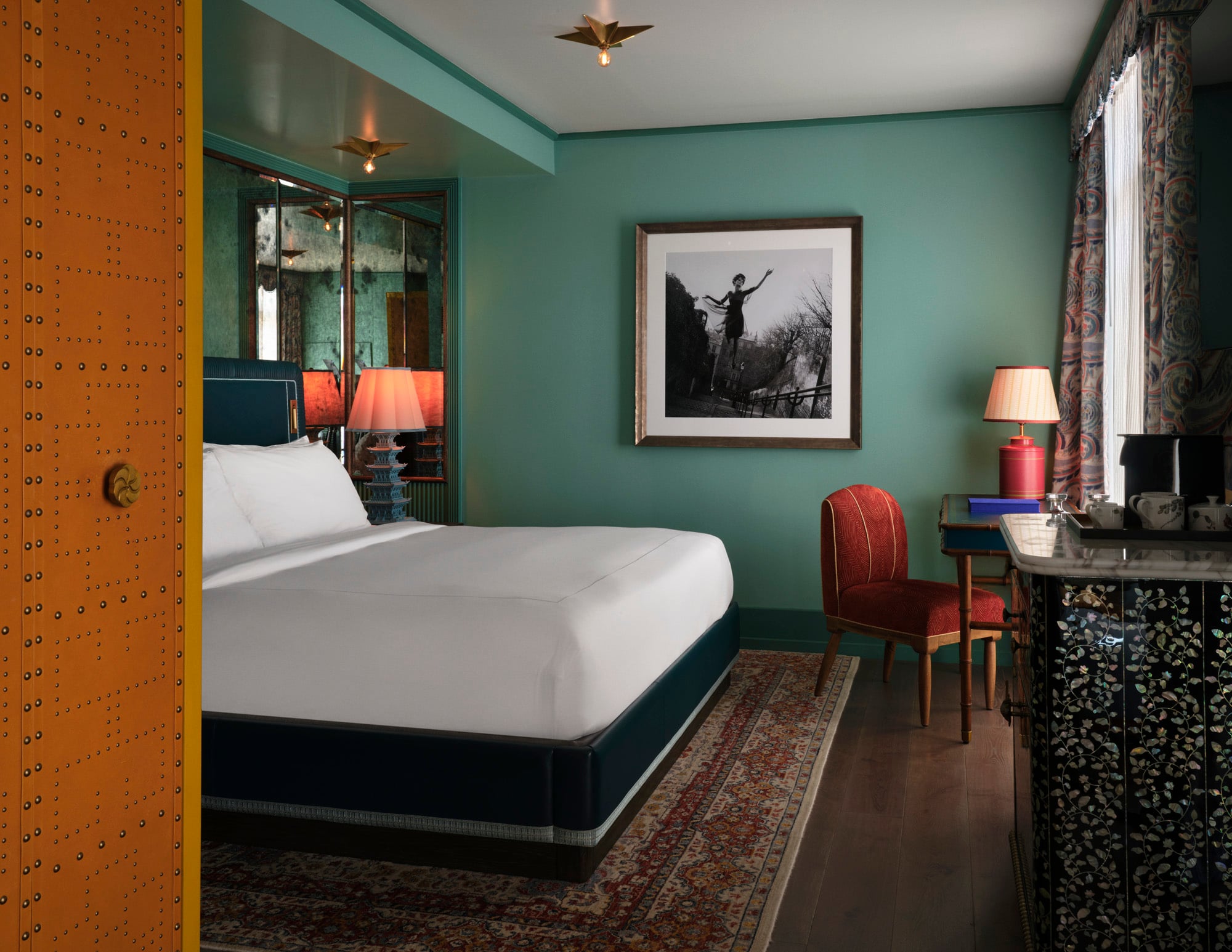 Hotel room featuring teal walls, an orange riveted door frame, a white bed, a framed black-and-white photo, and a red chair beside a glass-topped desk with a pink lampshade.