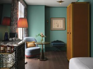 Teal-walled lounge corner with a round table, blue upholstered chairs, a violet bouquet in a glass vase, and a tall yellow riveted cabinet on the right. A framed sketch of eyes hangs on the wall near a small table.