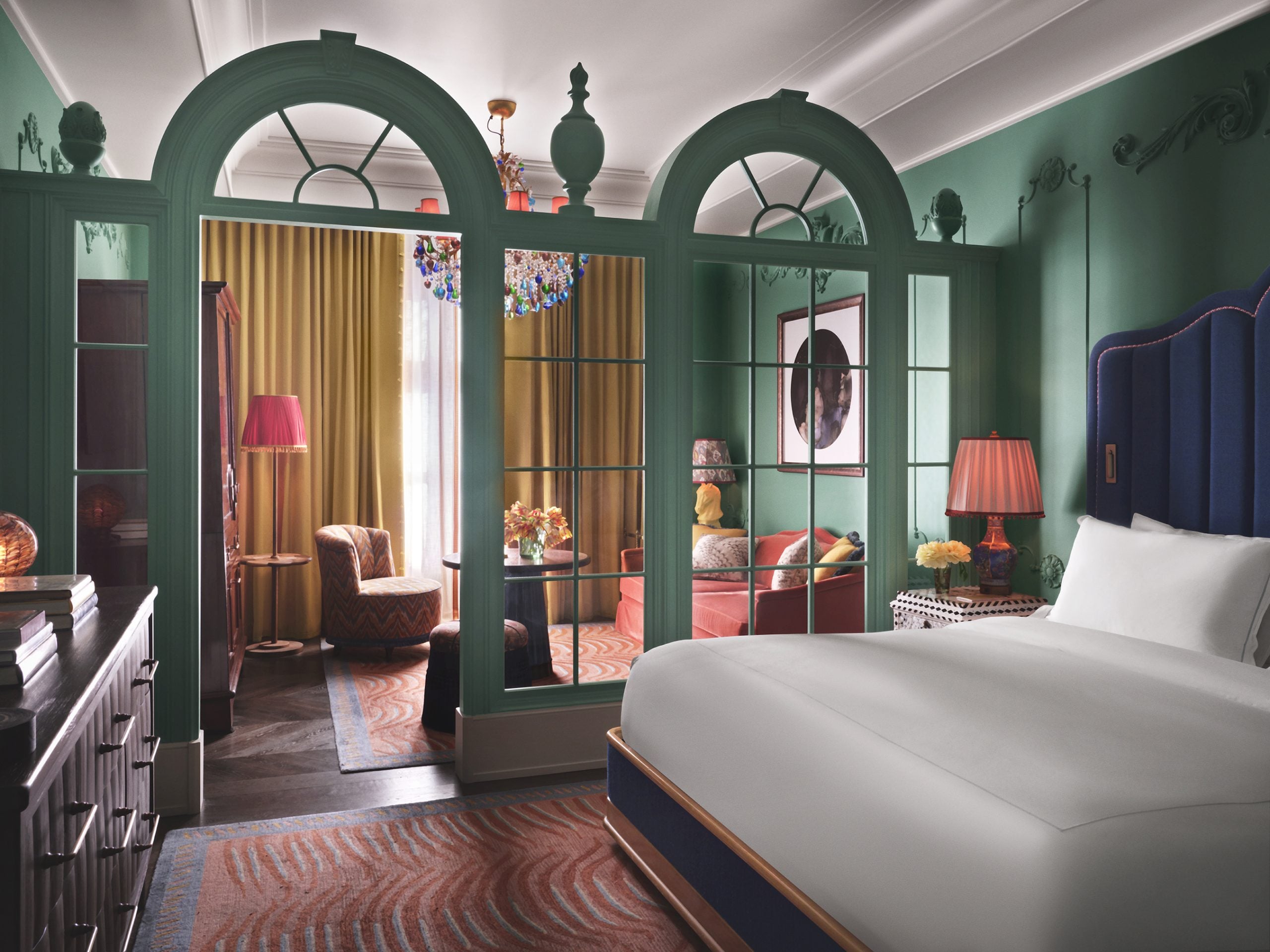 Hotel suite with green arched glass doors separating a pink-bed area from a living room, teal walls, and colorful lamps.