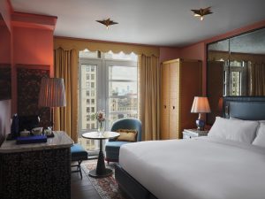 Hotel room with pink walls, blue chairs, a round glass table, and a large window showing city buildings outside.