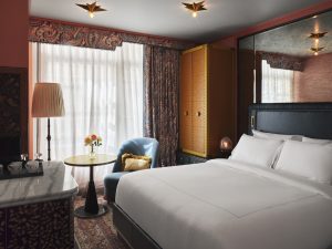 Hotel room with two large white beds, ornate wallpaper, a yellow safe cabinet, and a marble table with a vase of roses near a blue chair in front of a window.