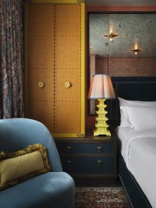 Hotel room corner with a tall yellow riveted cabinet next to a bed; a decorative yellow pagoda sits on a nightstand, blue chair in foreground.