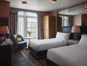 Hotel room with two white beds, a blue headboard, and patterned drapes beside a large window.