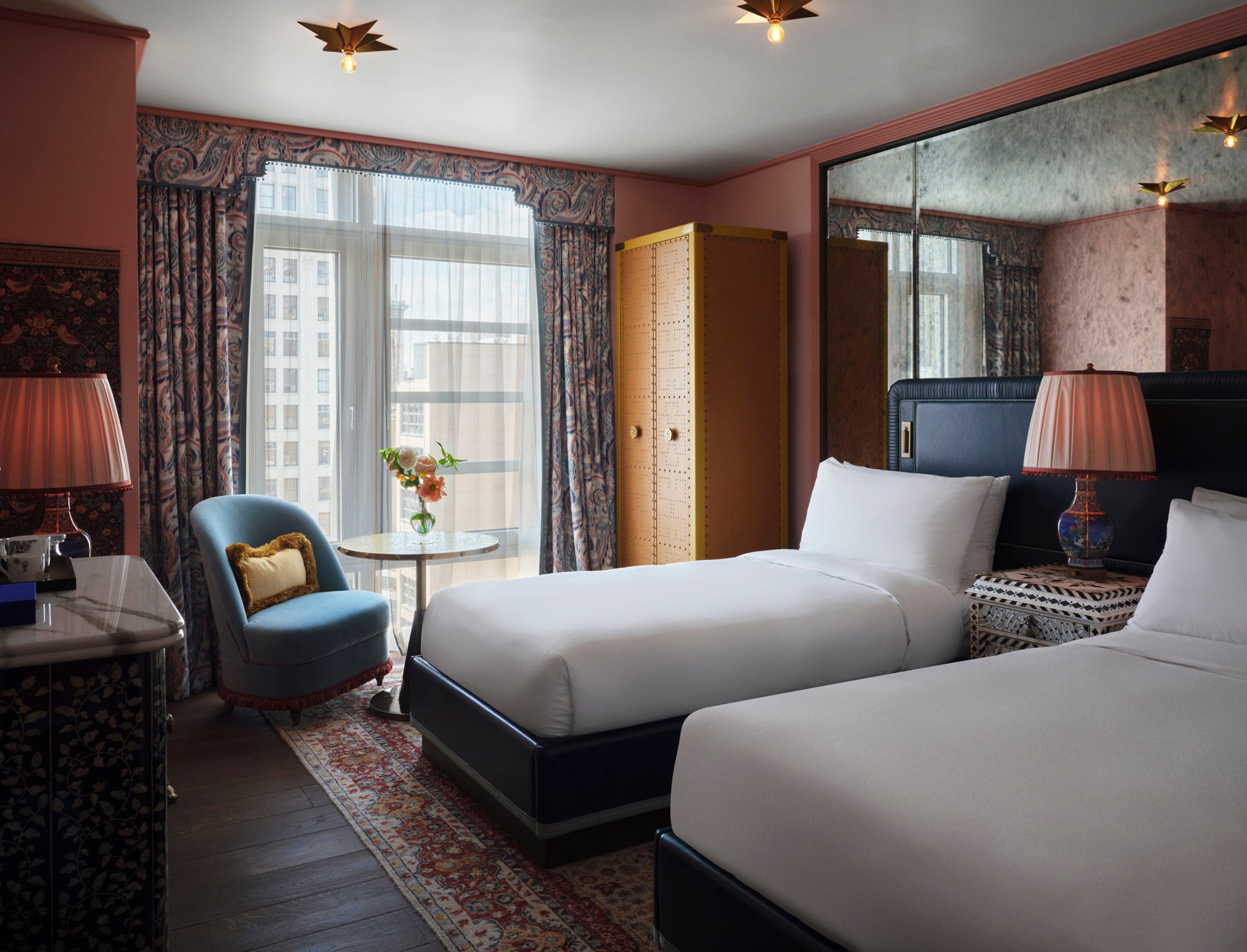 Hotel room with two white beds, a blue headboard, and patterned drapes beside a large window.