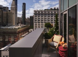 Rooftop balcony with wicker chairs and colorful cushions overlooking a city skyline with tall buildings and blue sky