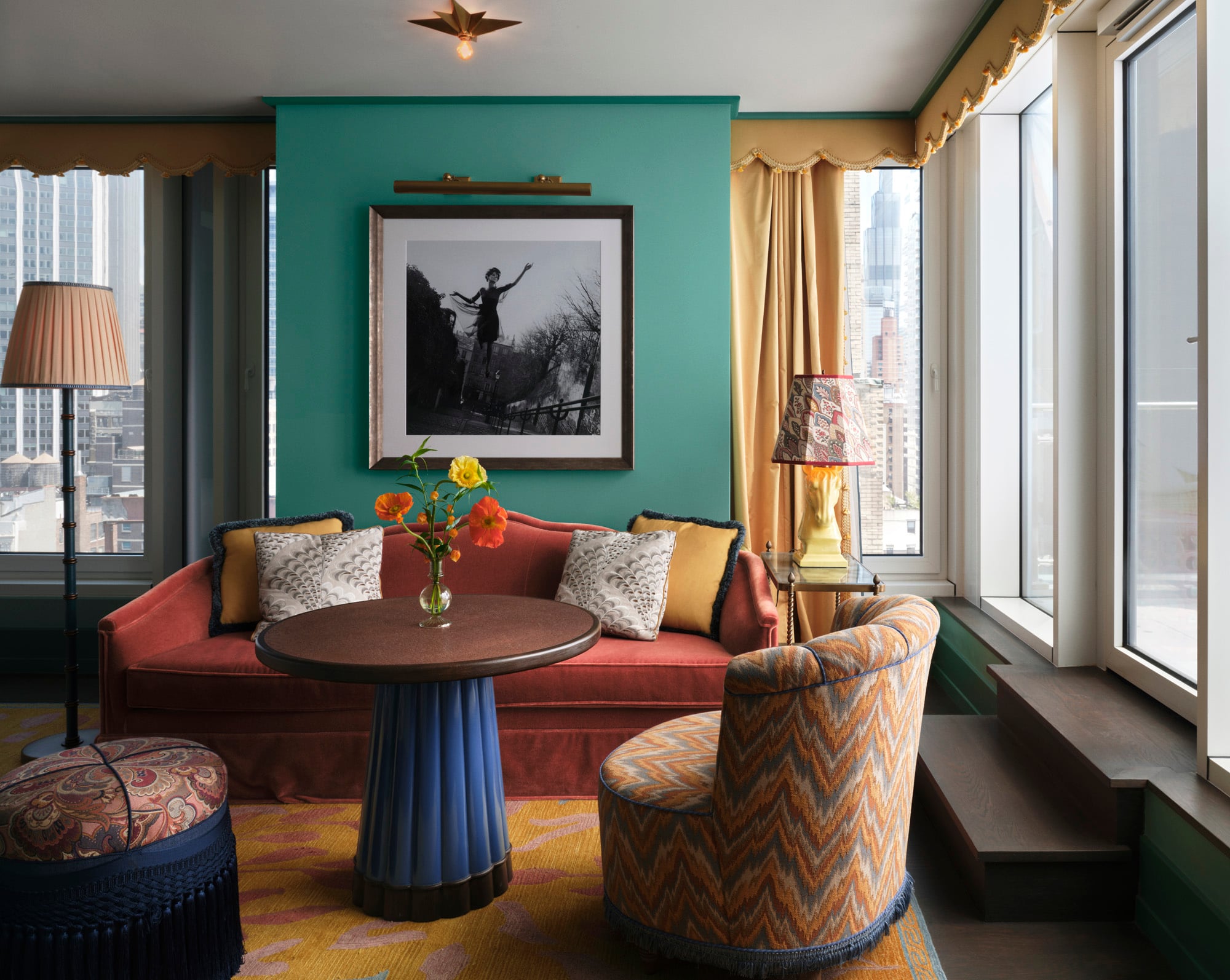 Teal accent wall with a framed black-and-white photo above an orange sofa; bright flowers on a round table in a sunlit living room.
