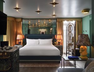 Luxurious hotel bedroom with a king bed, teal walls, gold curtains, and decorative screens. Warm pendant lights create a cozy glow.